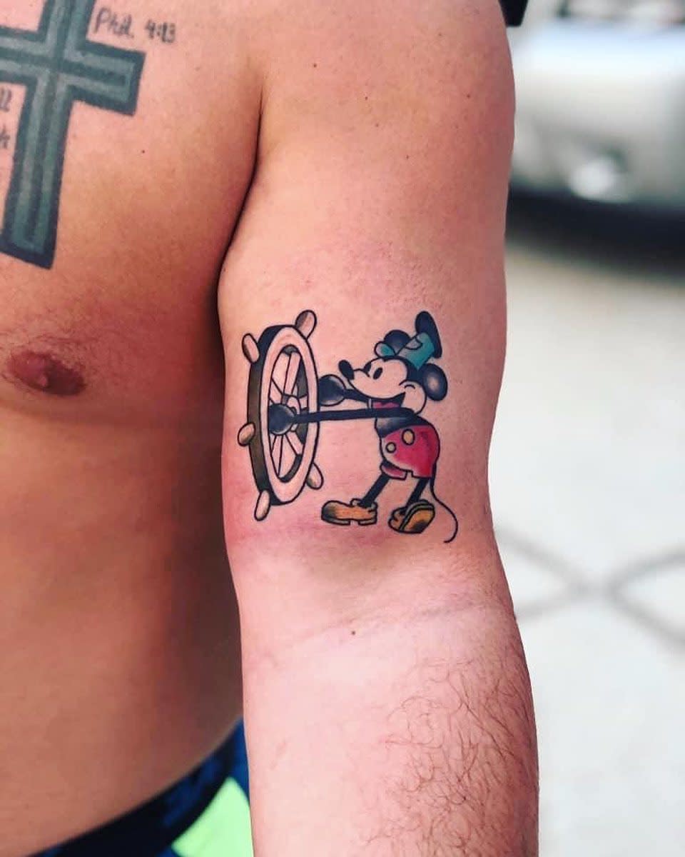Colorful tattoo of Steamboat Willie Mickey Mouse steering a ship's wheel, placed on the back of the arm